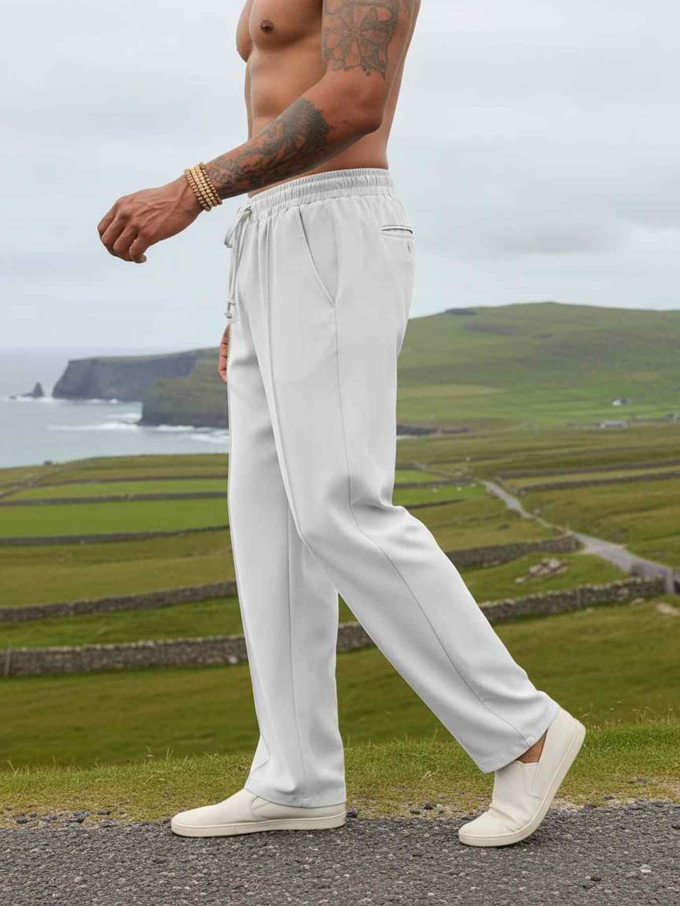 Men's light gray drawstring joggers with side pockets, worn outdoors on coastal path with green hills